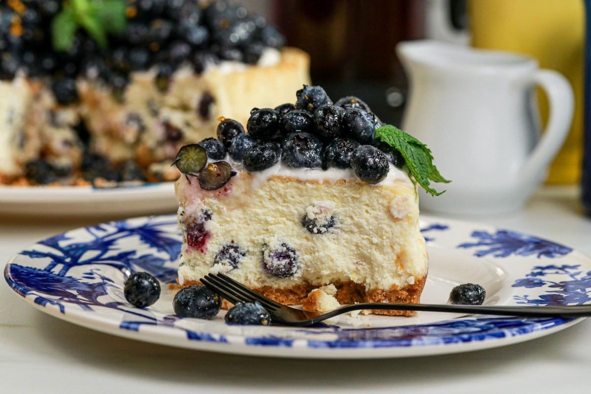 Gluten-free Lemon Blueberry Cheesecake slice in front of the sliced cake behind on blue white plate surrounded by lemon various cups and cutlery